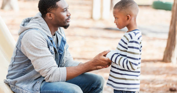 An African-American man in his 30s with a serious expression on his face, talking to his 7 year old son on a playground.